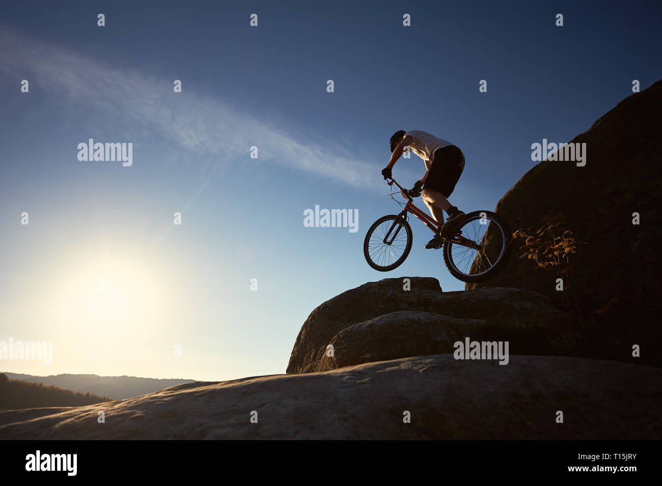 Professionista sportivo ciclista salto sulla bicicletta di prova sulla parte superiore del Rocky Mountain. Maschio rendendo motociclista acrobatico trucco in serata estiva, cielo blu e su Foto Stock