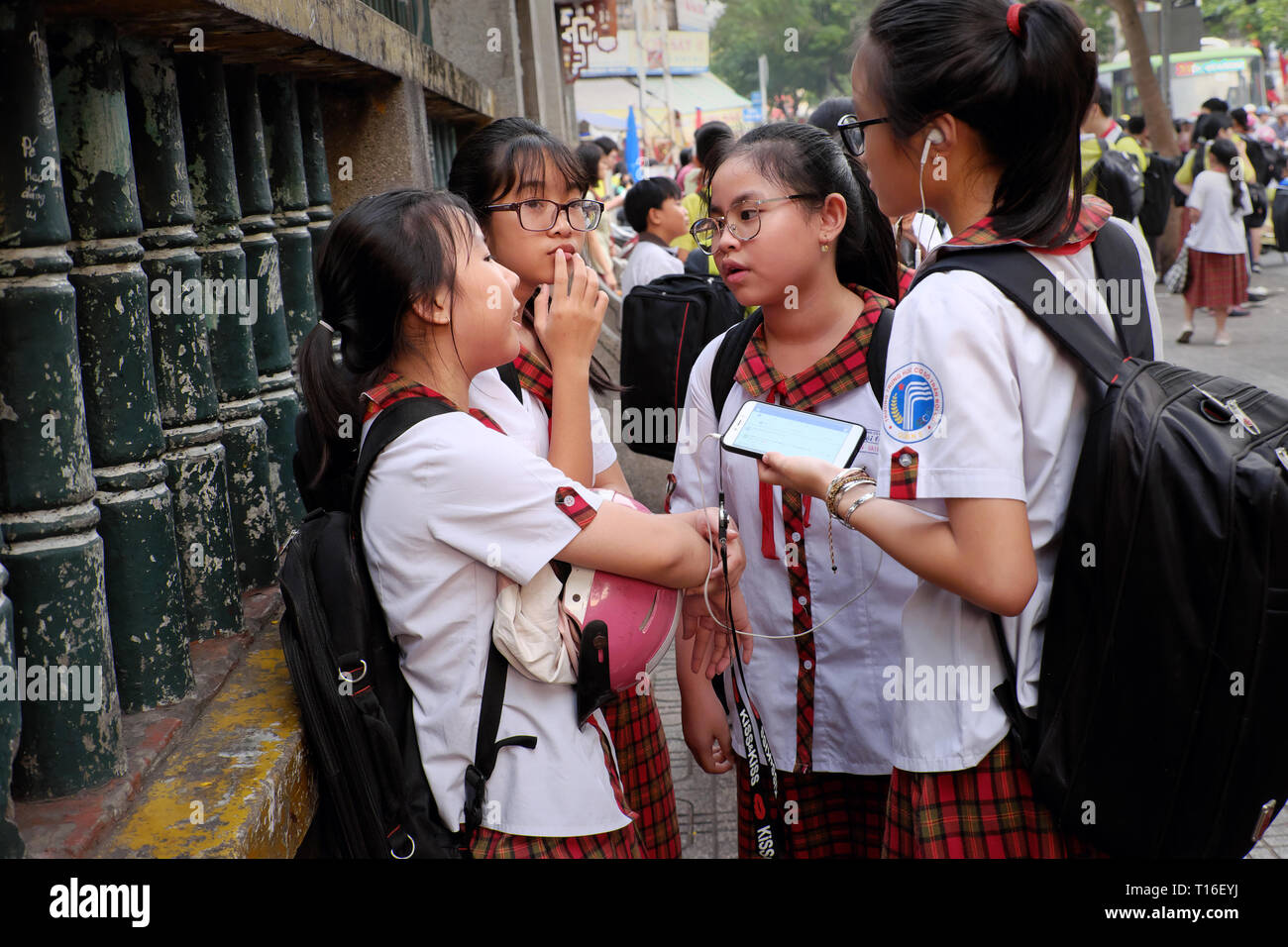 Gruppo di scuola ragazza di alzarsi e di parlare insieme a scuola gate, quattro ragazze adolescenti usura uniforme della pupilla, occhiali, una ragazza partecipare a smartphone, Vietnam Foto Stock