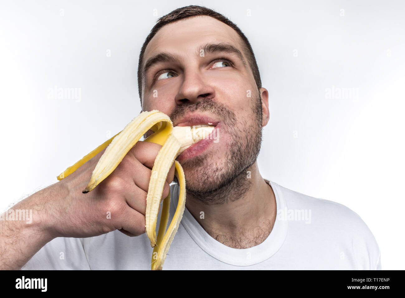 Un altro strano immagine di unshaved guy mangiare banane mature. Egli è mordere un grande pezzo di frutta. L uomo è godendo il mmoment e guardando da qualche parte a parte Foto Stock