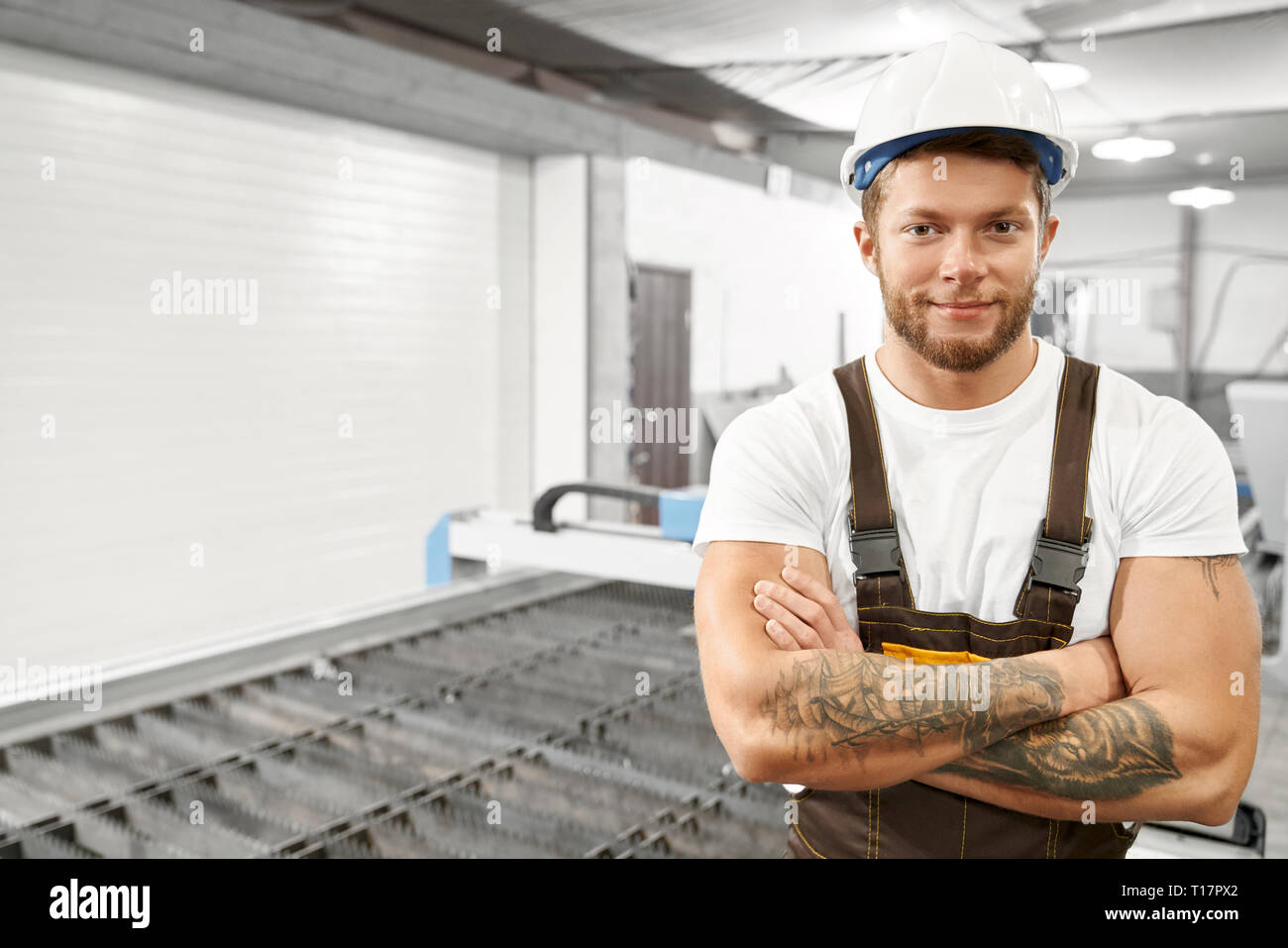 Ingegnere professionale e lavoratore di metallo in tute bianche, hardhat, stando in piedi in fabbrica. Bello, brutale uomo con tatuaggi in posa con mani incrociate, guardando la fotocamera e sorridente. Foto Stock