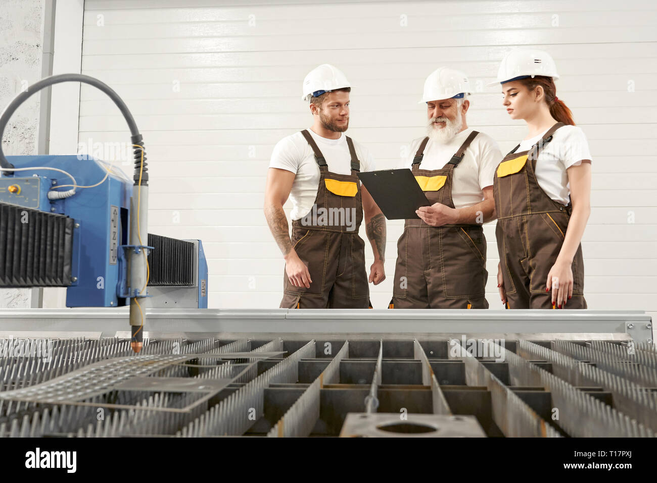 CNC laser plasma cutter in fabbrica metallurgica. Tre operatori professionali in piedi, parlando, guardando alla cartella. Gli ingegneri di indossare tute e caschi bianco e magliette. Foto Stock