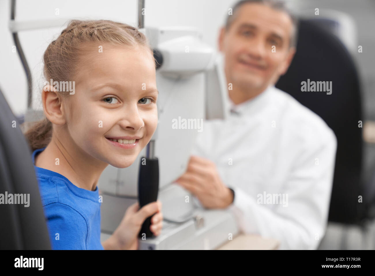 Paziente esame della vista ottico in ufficio. Bella ragazza adolescente in posa, sorridente e guardando la fotocamera. Oculista e lampada a fessura macchina su sfondo. Foto Stock