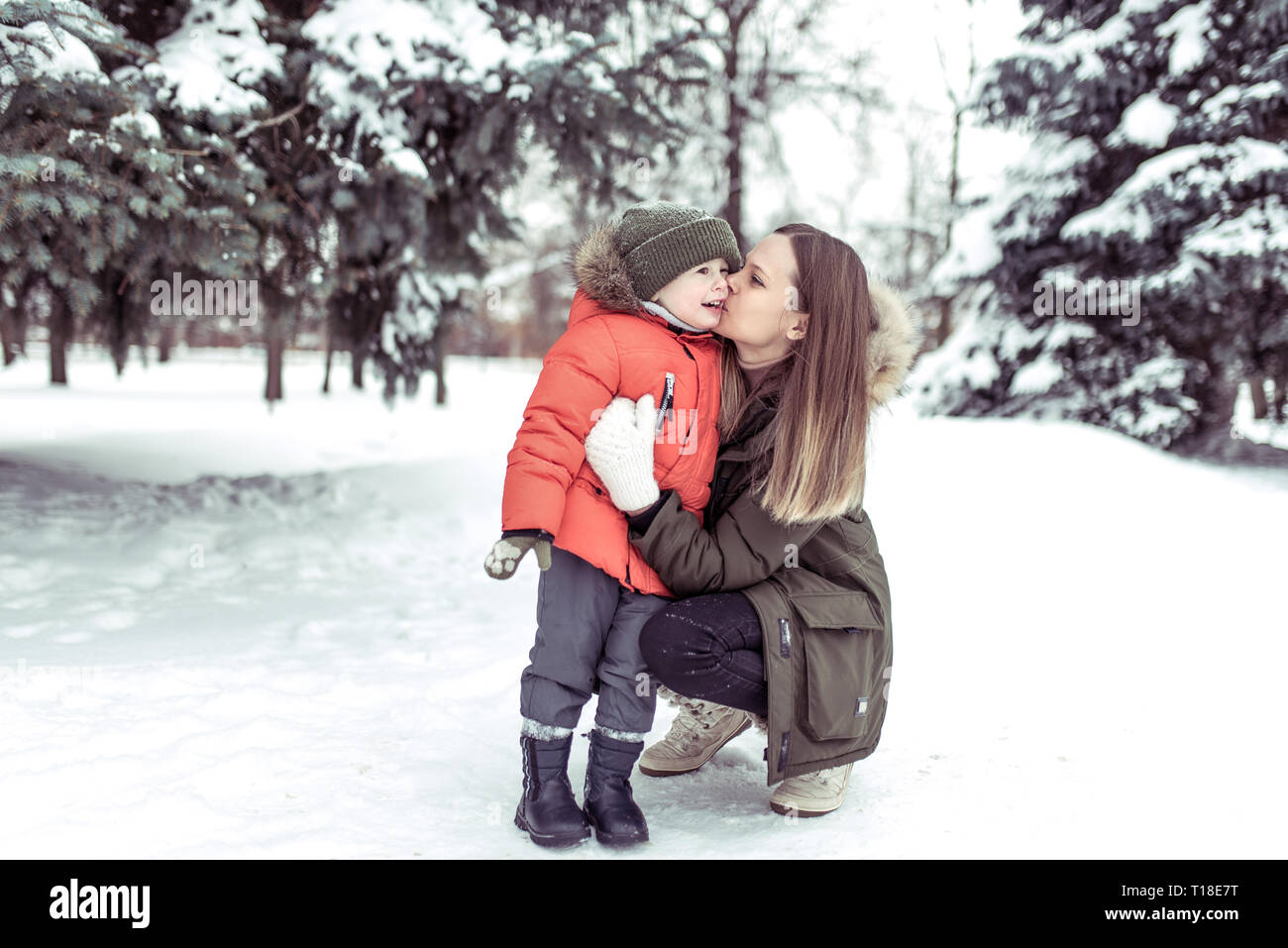 Una giovane madre baciare il suo bambino, ragazzo di 3-5 anni, in inverno nella foresta, contro uno sfondo derive di neve coperto di neve e gli alberi di Natale. Emozioni Foto Stock