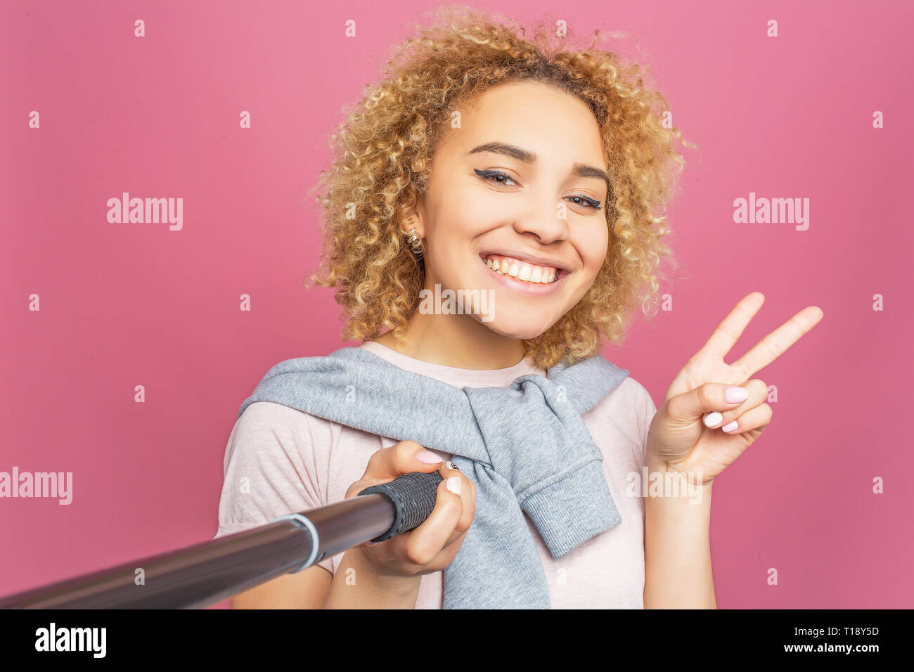 Sorridente ragazza sta prendendo un selfie. Essa utilizza un selfie sticj per l. Giovane donna è in posa e mostrando il pezzo simbolo per telecamera. Isolato sul retro di colore rosa Foto Stock