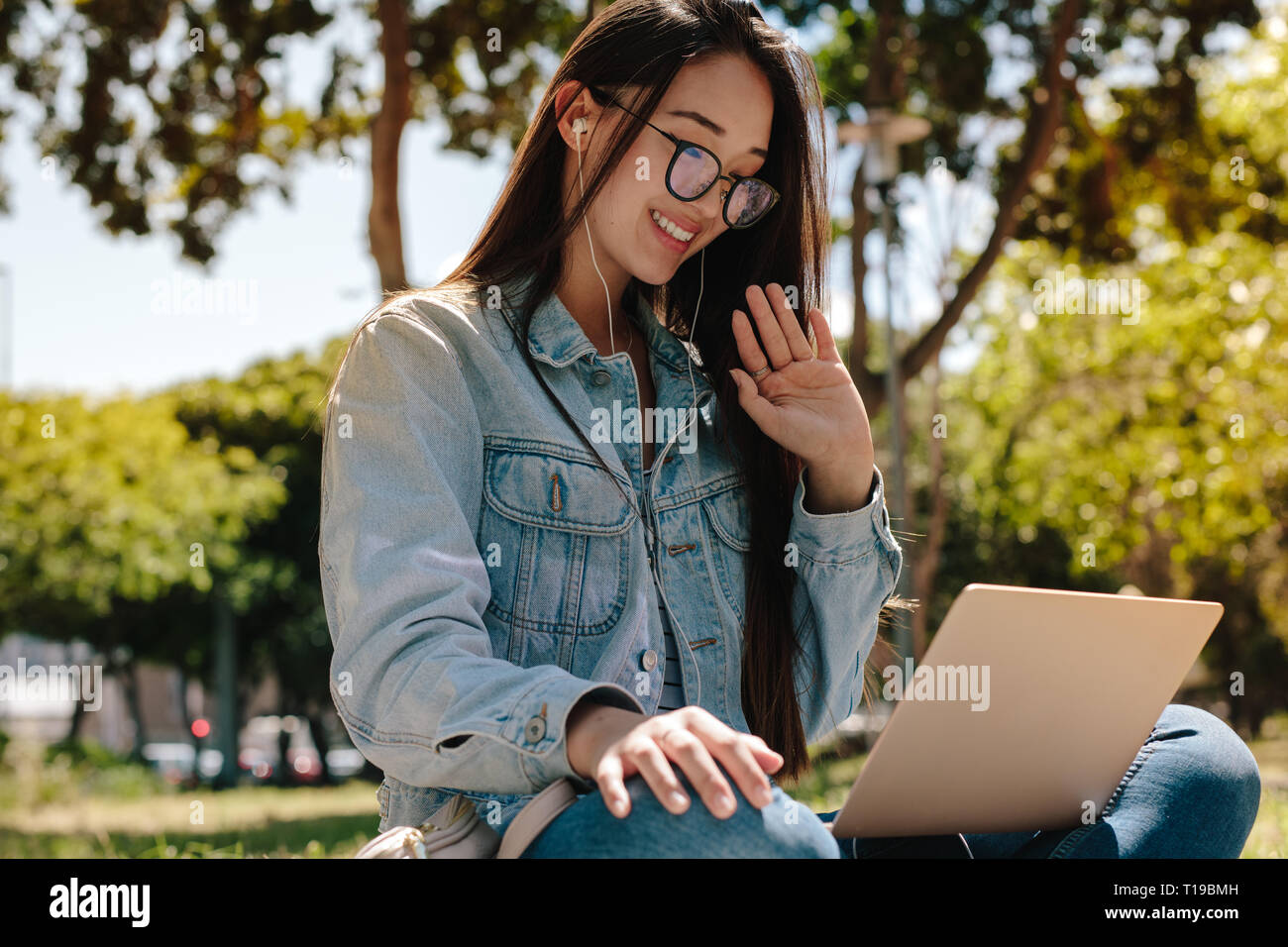Close up di un college girl su una chiamata video seduto in un parco. Ragazza asiatica facendo una chat video con un computer portatile e di auricolari seduti all'aperto. Foto Stock