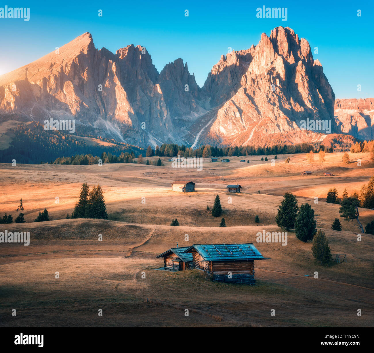 Piccole case in legno nella bella valle di montagna al tramonto. In autunno il paesaggio con le montagne, edifici, collina con erba gialla, alberi, rocce alte, sky Foto Stock Piccole case in legno nella bella valle di montagna al tramonto. In autunno il paesaggio con le montagne, edifici, collina con erba gialla, alberi, rocce alte, sky Foto Stock