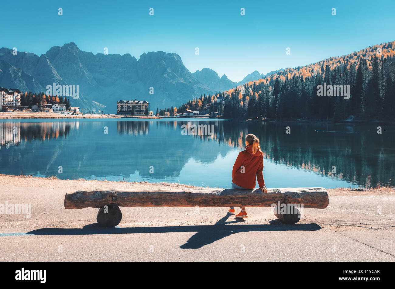 Giovane donna seduta sul banco di legno sulla costa del lago di Misurina al tramonto in autunno. Dolomiti, Italia. Paesaggio con ragazza in giacca rossa, reflec Foto Stock