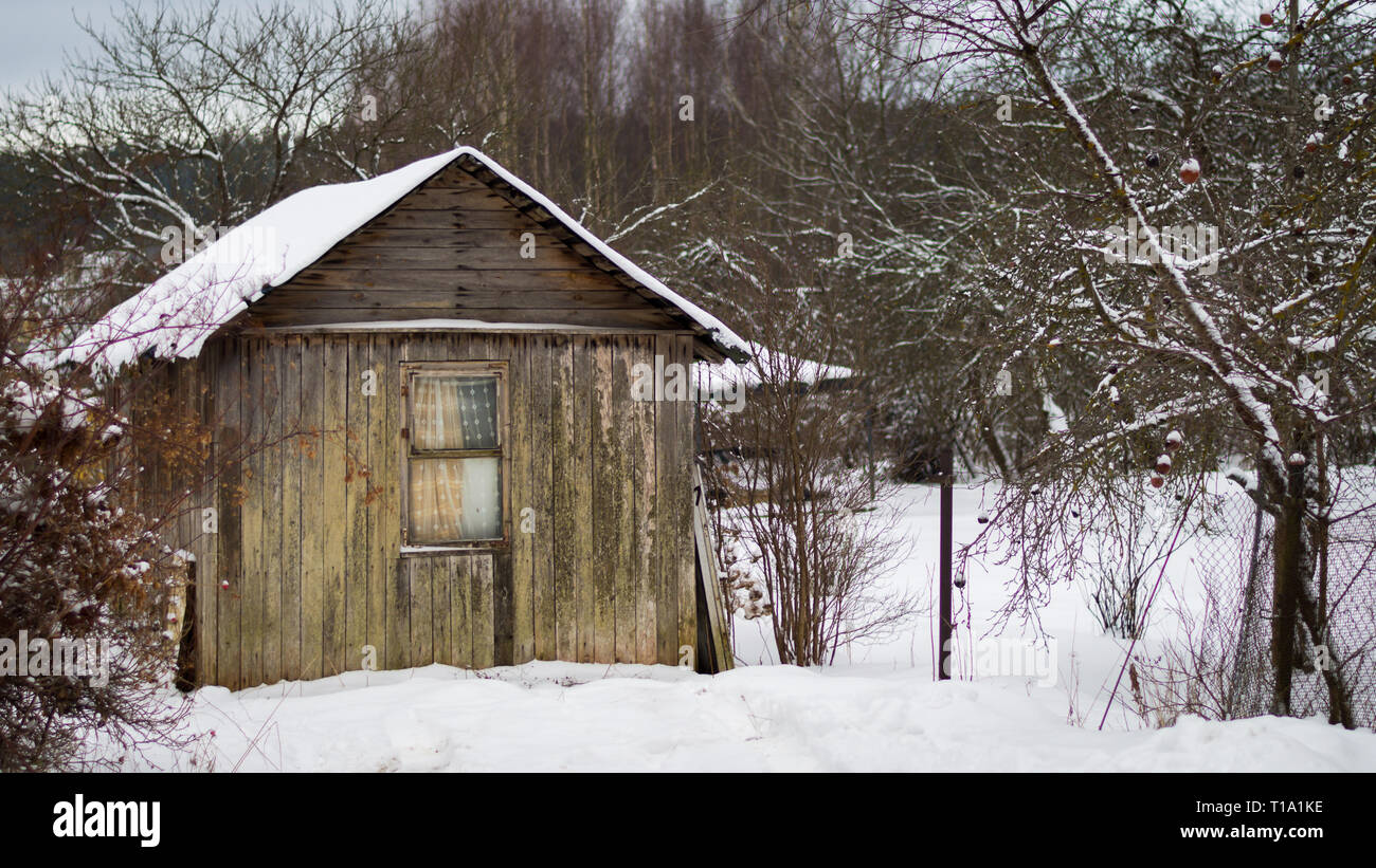Una piccola vecchia casa in legno con una finestra, una recinzione e un piccolo giardino in inverno coperto di neve Foto Stock
