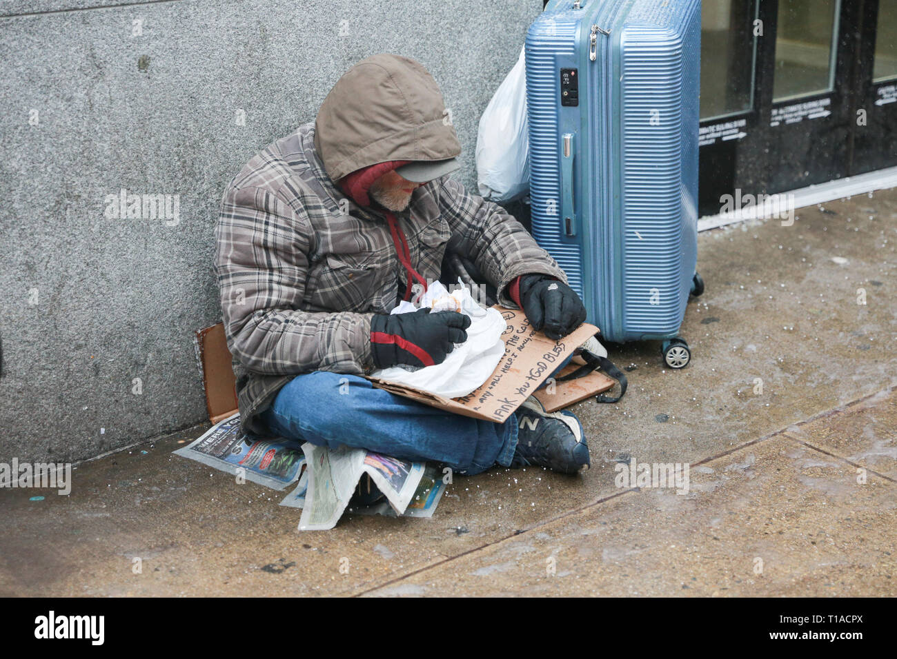 New York, NY - 12 Febbraio 2019: un senzatetto è mangiare cibo, mentre tenendo un cartello per aiutare, cibo e denaro. Midtown Manhattan. - Immagine Foto Stock