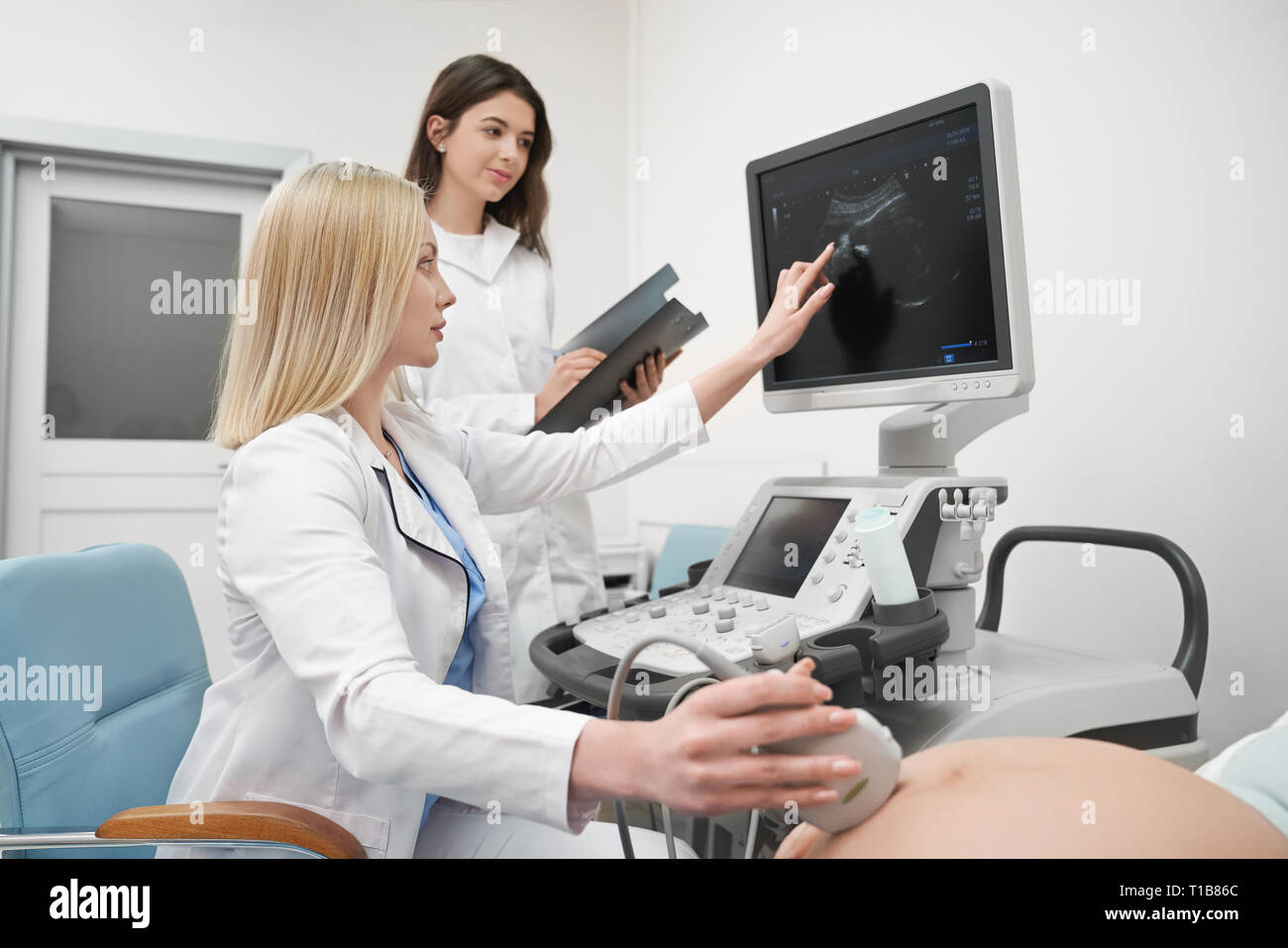 Terapista esaminando l'addome della donna gravida. Medico professionale dispositivo di trattenimento, guardando il monitor della macchina ad ultrasuoni. L'infermiera aiutando, apposita cartella, lavorando in clinica medica. Foto Stock