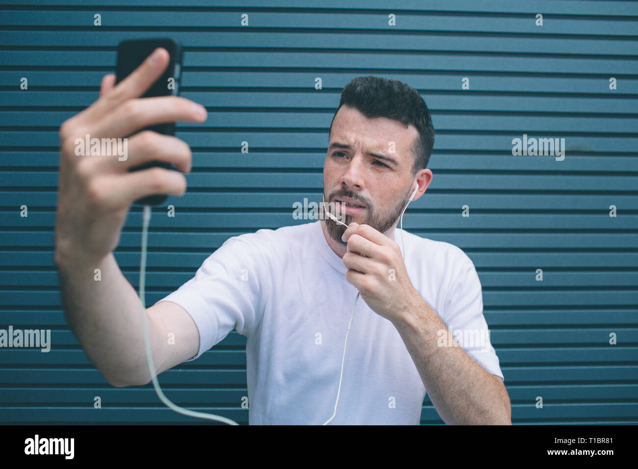Il ragazzo è in posa sulla fotocamera. Egli è tenuto selfie e tenendo una mano sul mento. Anche l uomo è l'ascolto di musica. Isolato su striped e sfondo blu. Foto Stock