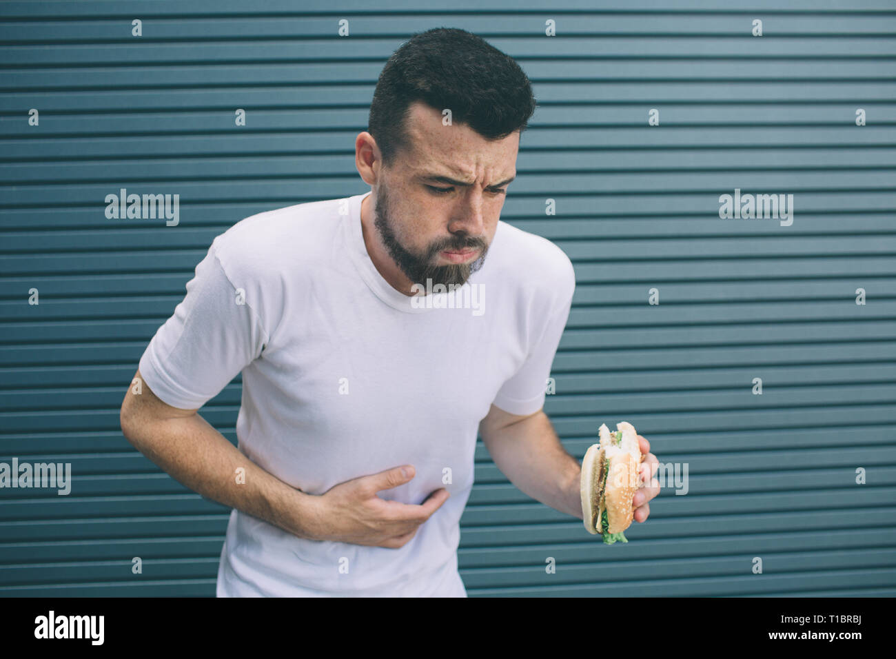 Guy è malato. Egli è in possesso di burger in mano e guardando verso il basso. L uomo è di andare a vomitare. Isolato su striped e sfondo blu. Foto Stock