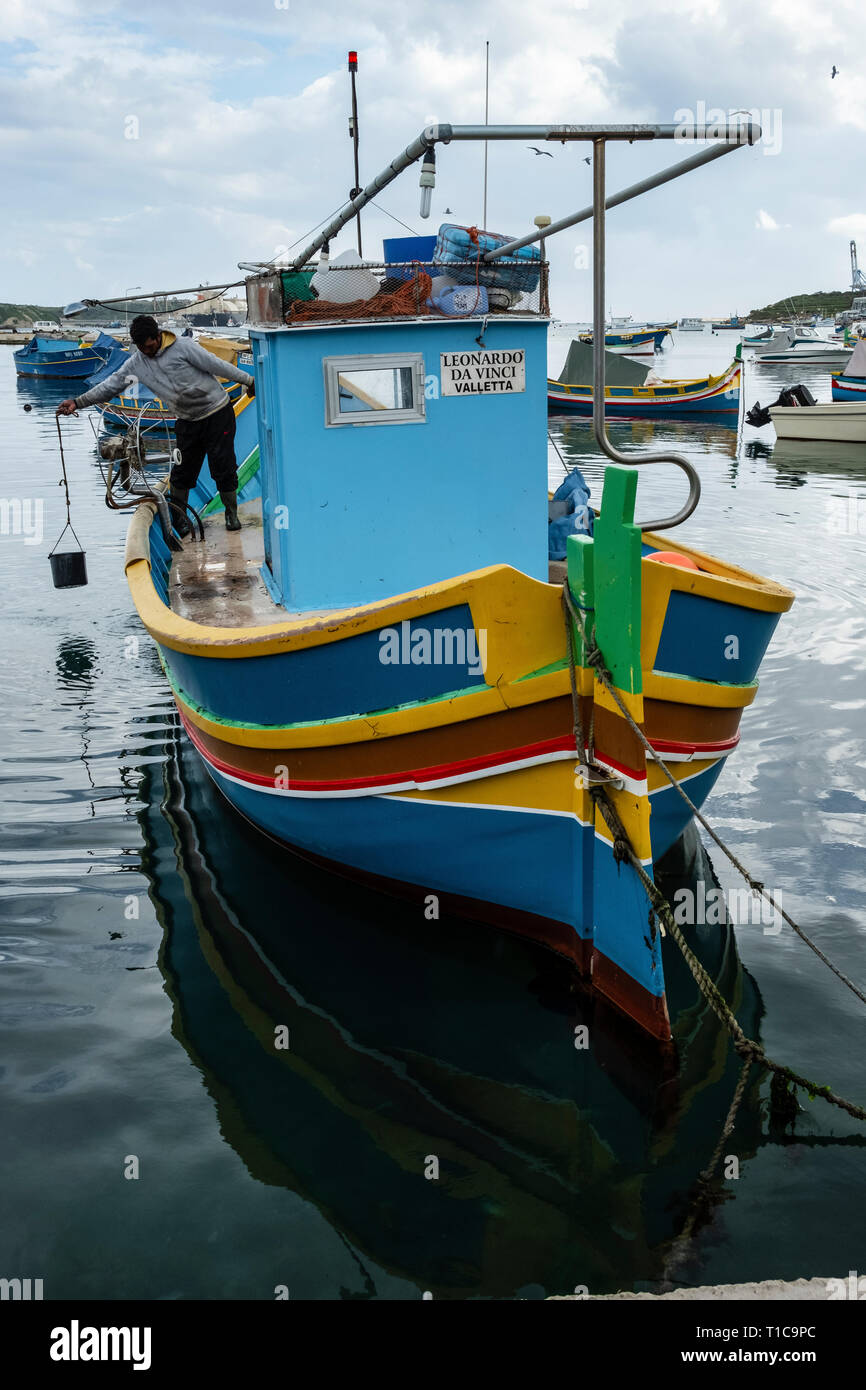 Un pescatore di raccolta di acqua per lavare il ponte di una tradizionale maltese colorate barche da pesca (luzzu), Marsaxlokk, Malta Foto Stock