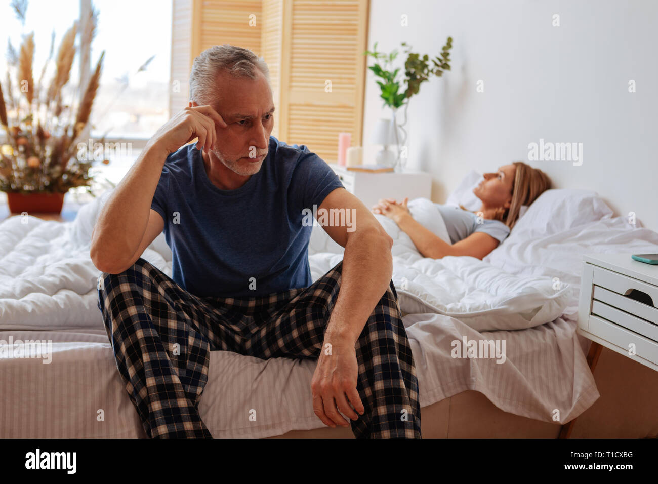 Grey-haired marito indossando pigiami seduto vicino a dormire moglie Foto Stock