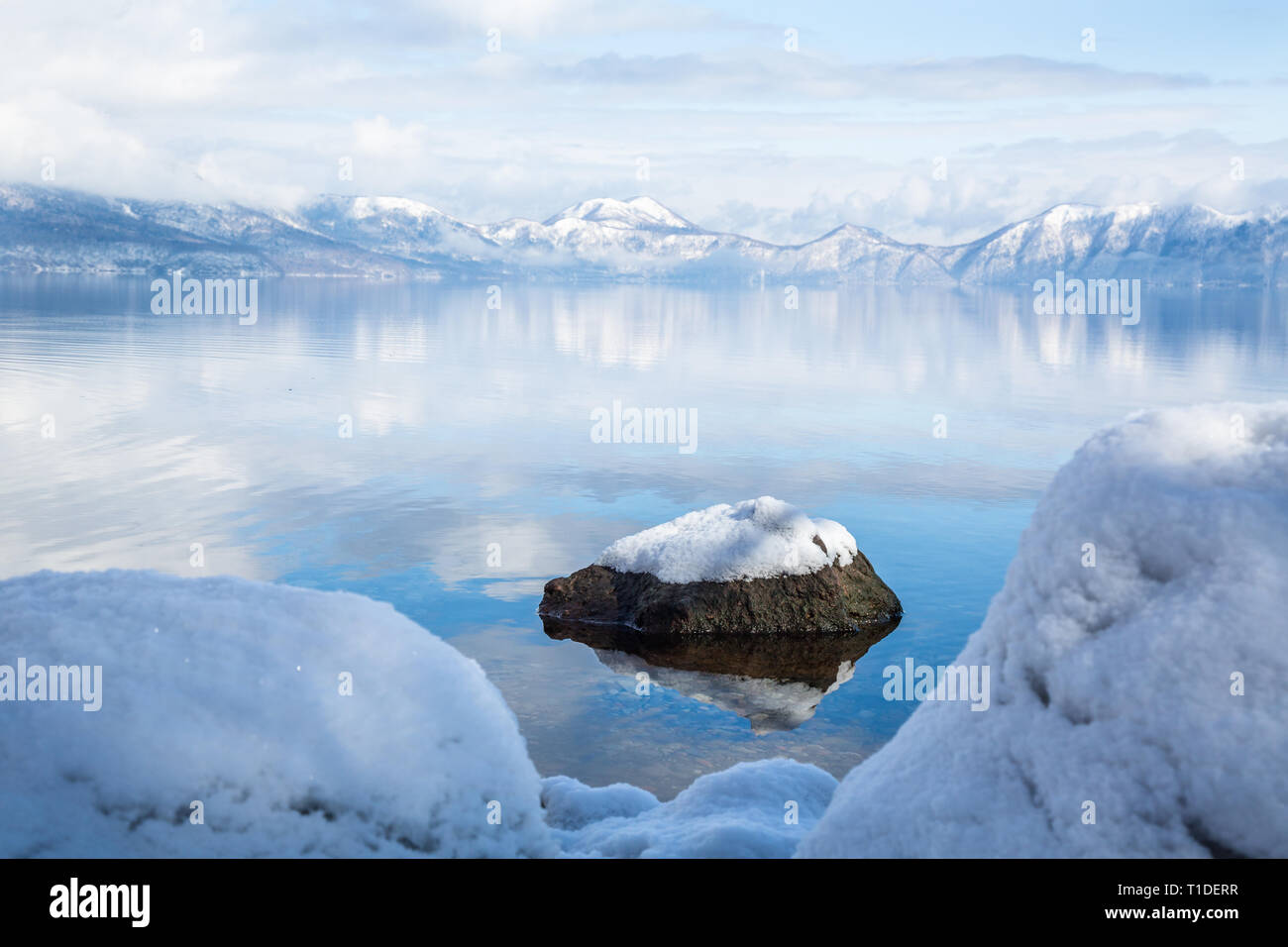 Acqua calma nel lago di Shikotsu, Giappone riflette le montagne in lontananza e rocce sul litorale sono coperti di neve fresca. Foto Stock