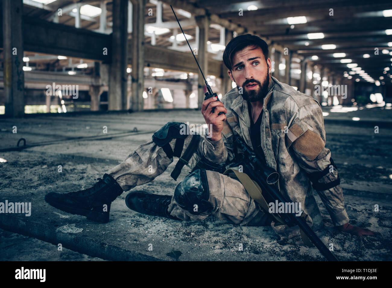 Bella immagine di attraente e bella bruna seduto per terra. Egli è in possesso di radio portatile nella mano sinistra. Fucile è giacente sulla sua gamba sinistra. Guy Foto Stock