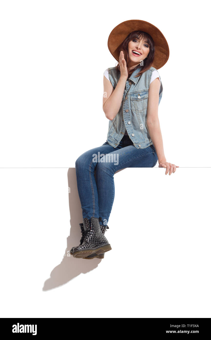 Giovane donna in jeans giubbotto, stivali neri e pelle scamosciata marrone cappello è seduta su una parte superiore con le gambe incrociate a caviglia, guardando la fotocamera e ridere. A piena lunghezza Foto Stock