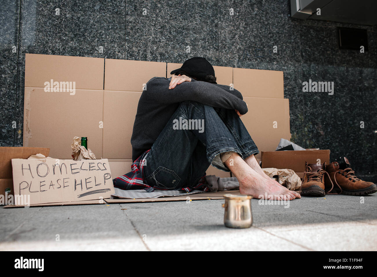 Una foto del disperato uomo seduto sul cartone e a nascondere il suo volto. Egli è coperto con le mani e guardando verso il basso. L uomo è seduta senza scarpe Foto Stock