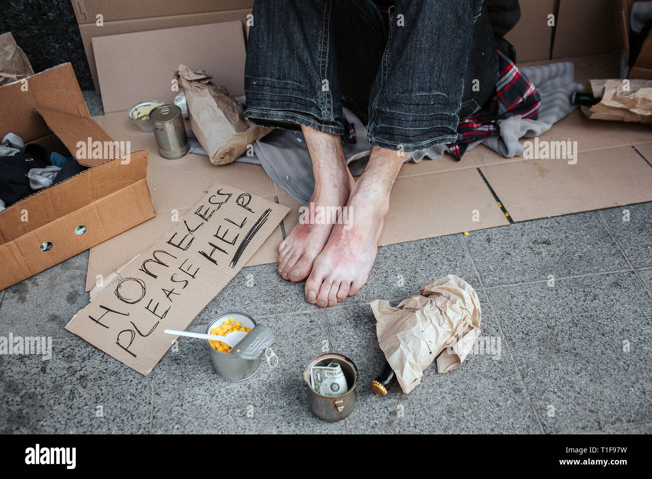 Una immagine di uomo di piedi senza scarpe. L uomo è seduto sul cartone. Egli è un mendicante. Vi è un cartello che dice di senzatetto si prega di aiutare, bicchiere di metallo con Foto Stock