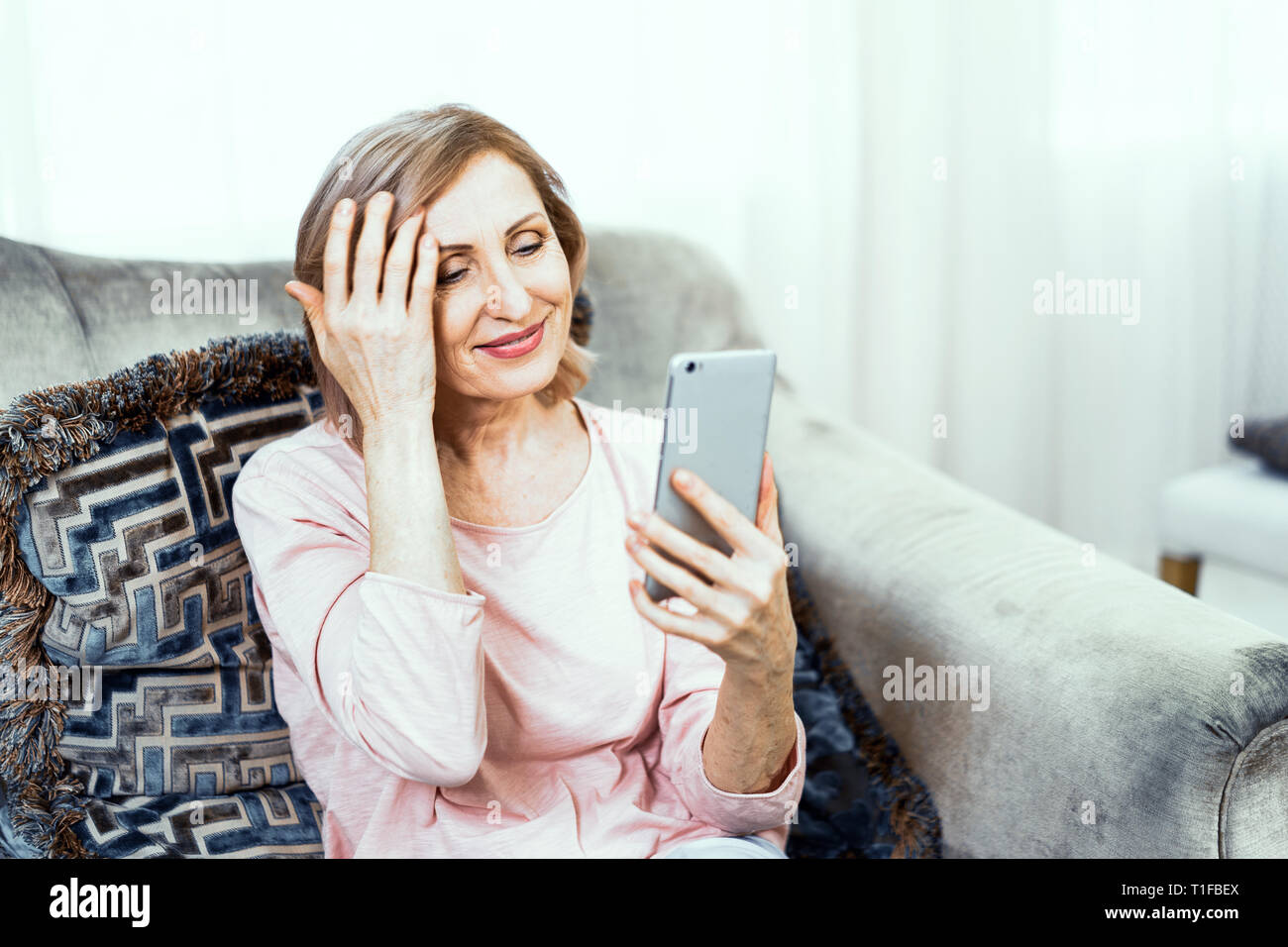 Donna anziana con un telefono nelle sue mani di buon umore è in appoggio in casa nel salotto. Foto Stock