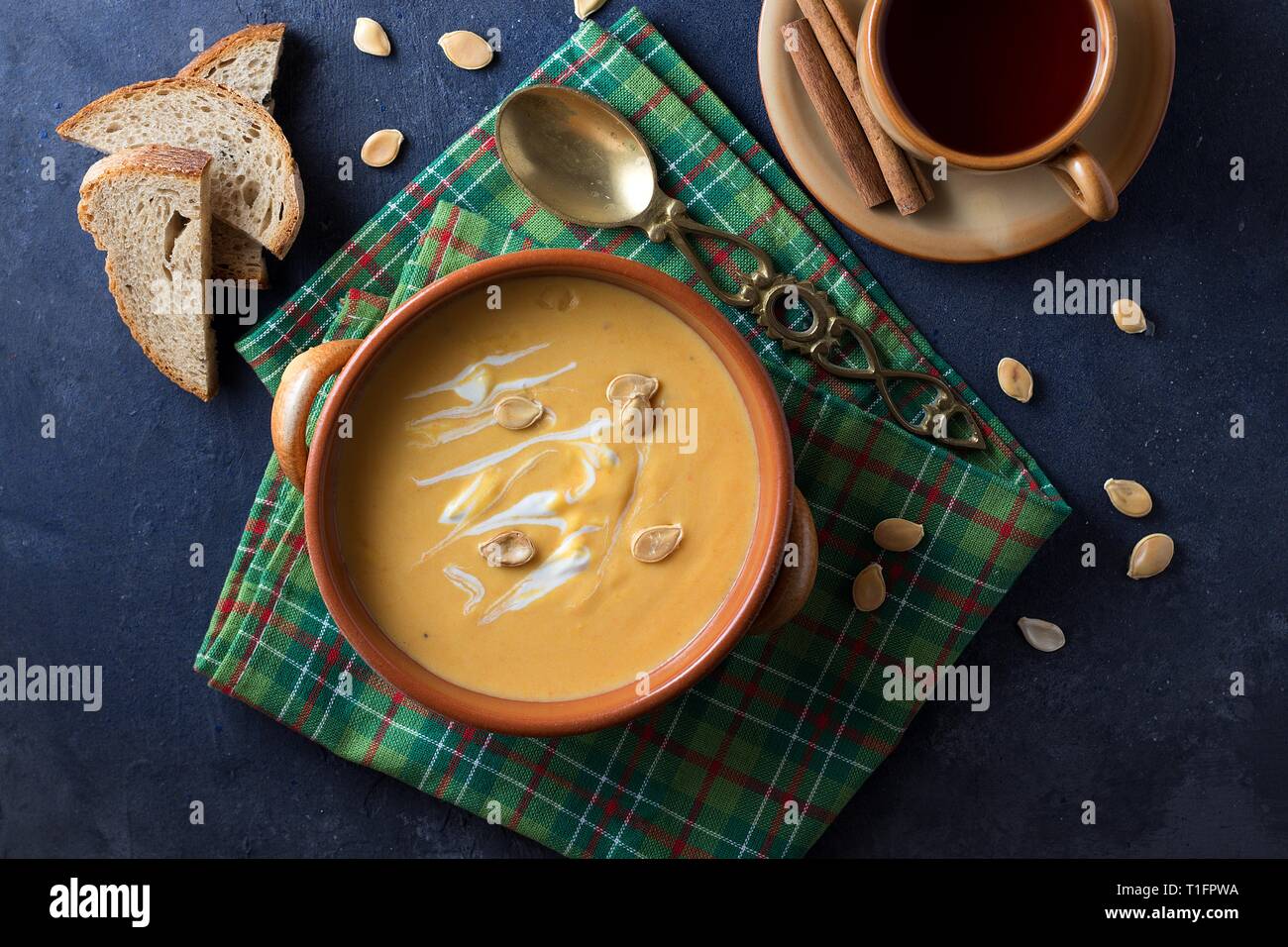 Vista dall'alto in basso sul vaso di terracotta di zuppa di zucca con crema su sfondo nero, tavolo verde asciugamano cucchiaio in ottone e la tazza di tè di frutta Foto Stock