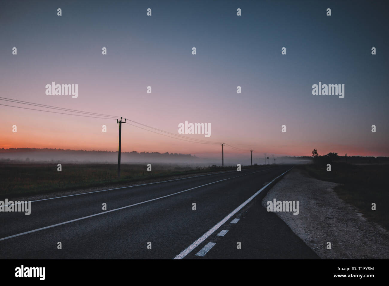Mattina è venuto. Il sole arancione aumenta al di sopra di un campo di misty, strada asfaltata e la foresta di alberi scuri. Foto Stock