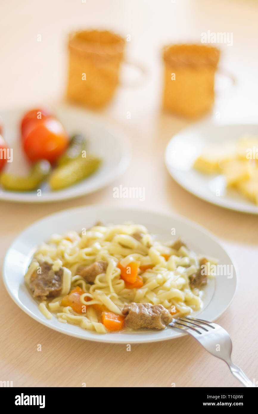 Pasta con le fette di carne e carote. Marinata di pomodori e cetrioli. Formaggio a fette in una piastra. Due tazze sul tavolo. Foto Stock