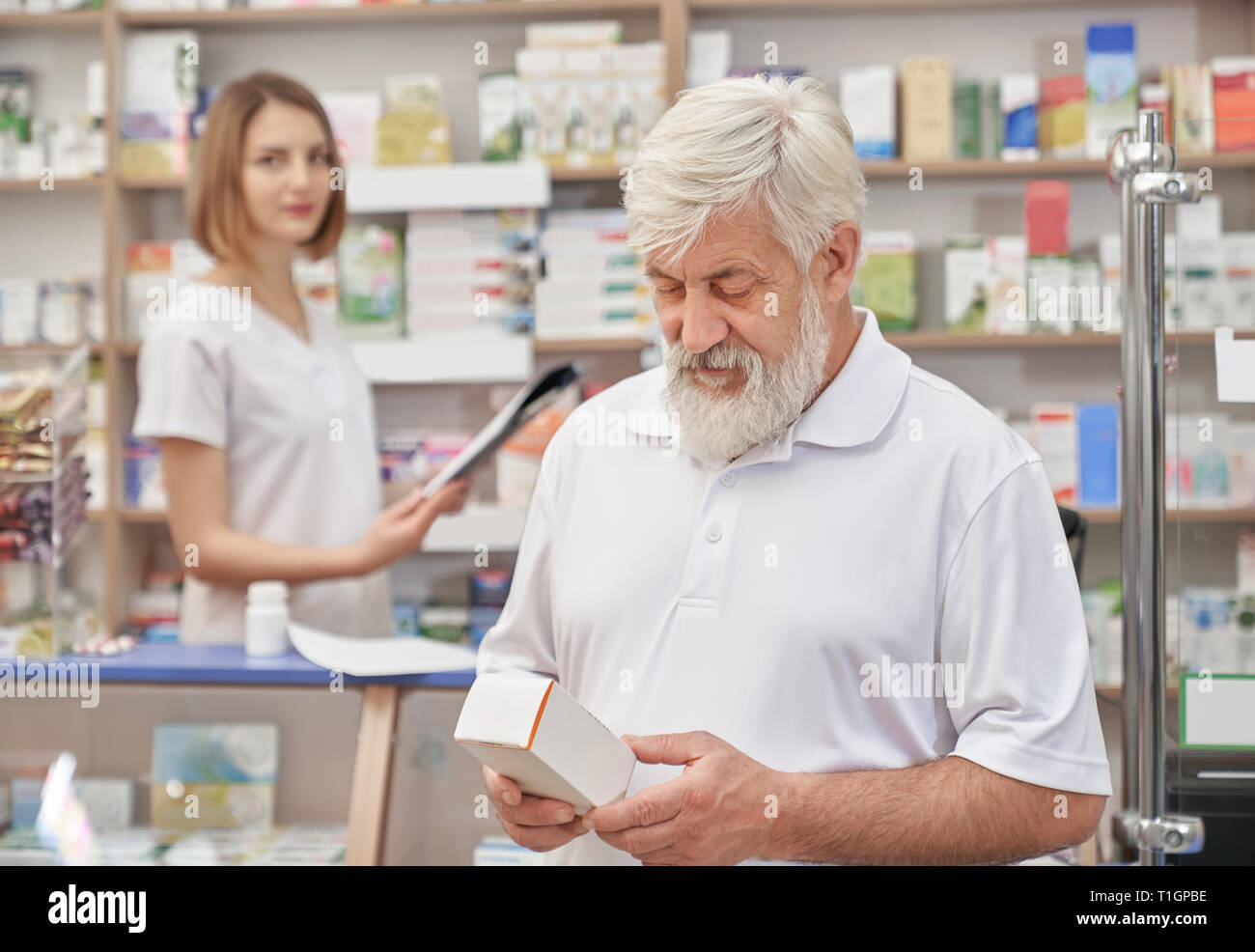 Uomo anziano scegliendo i medicamenti in farmacia, pensando. Titolare di pensione o di rendita barbuto in bianco t shirt azienda cassetta medica, guardando verso il basso. Il farmacista femmina permanente al contatore in farmacia. Foto Stock