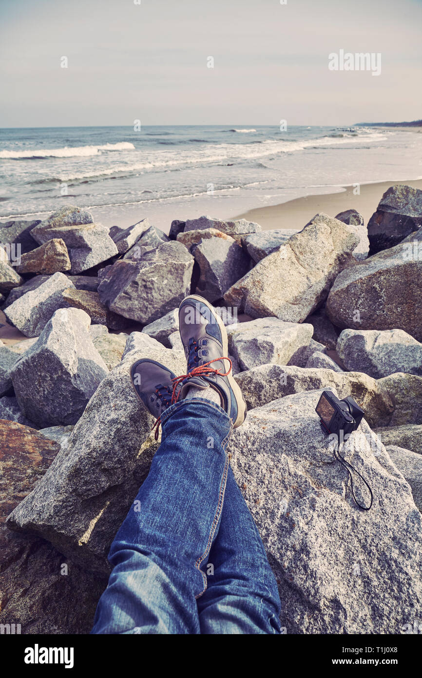 Le gambe di una persona a riposo su una spiaggia rocciosa con una telecamera a parte, dai toni di colore foto, messa a fuoco selettiva. Foto Stock