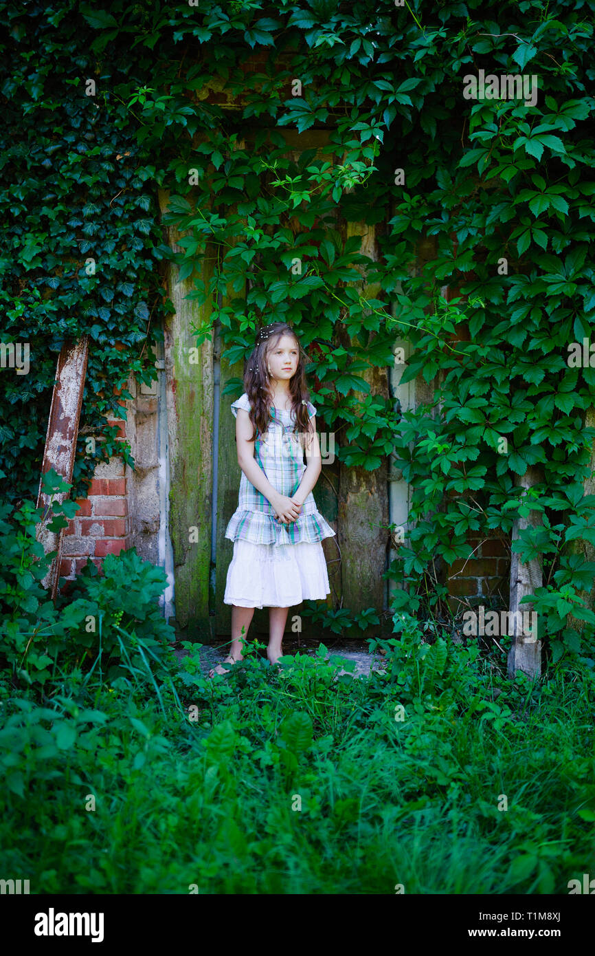 Ragazza serena in abito in piedi alla porta rustica coperta di edera verde Foto Stock