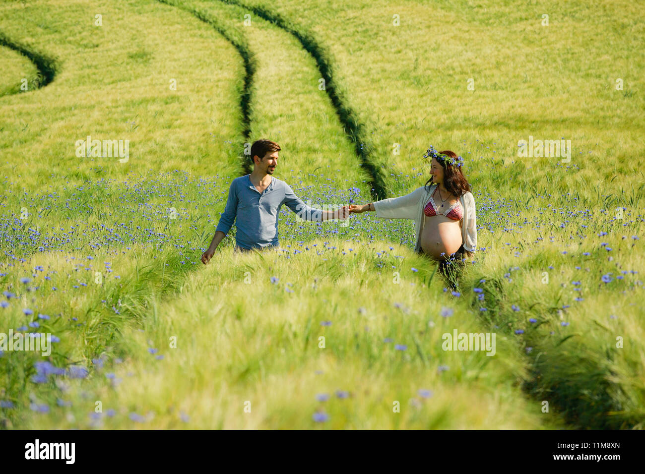 Affettuosa coppia incinta che tiene le mani, camminando nel soleggiato, idilliaco campo rurale verde Foto Stock