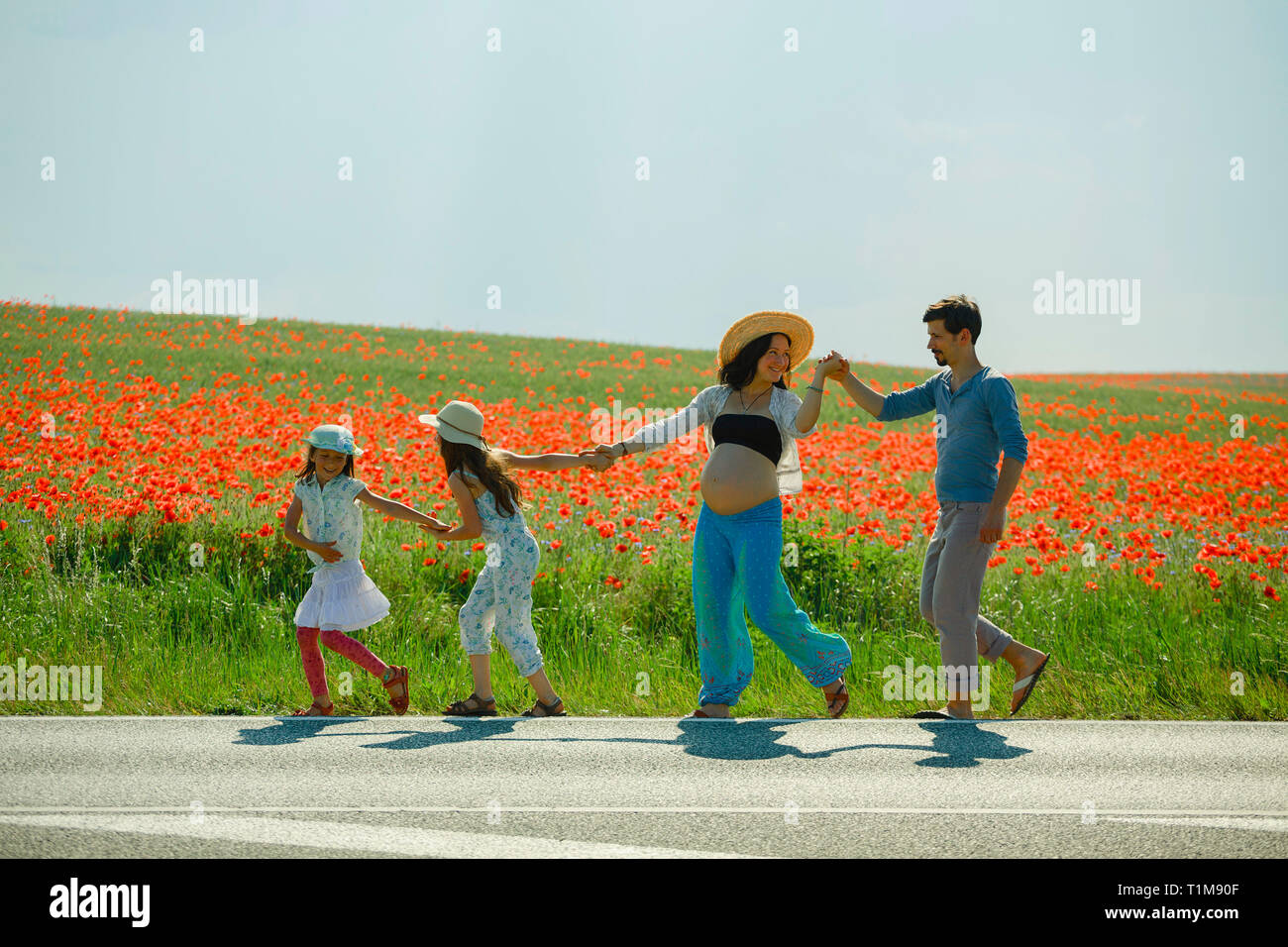 Felice famiglia incinta che tiene le mani, camminando lungo il soleggiato campo di papavero rurale rosso Foto Stock