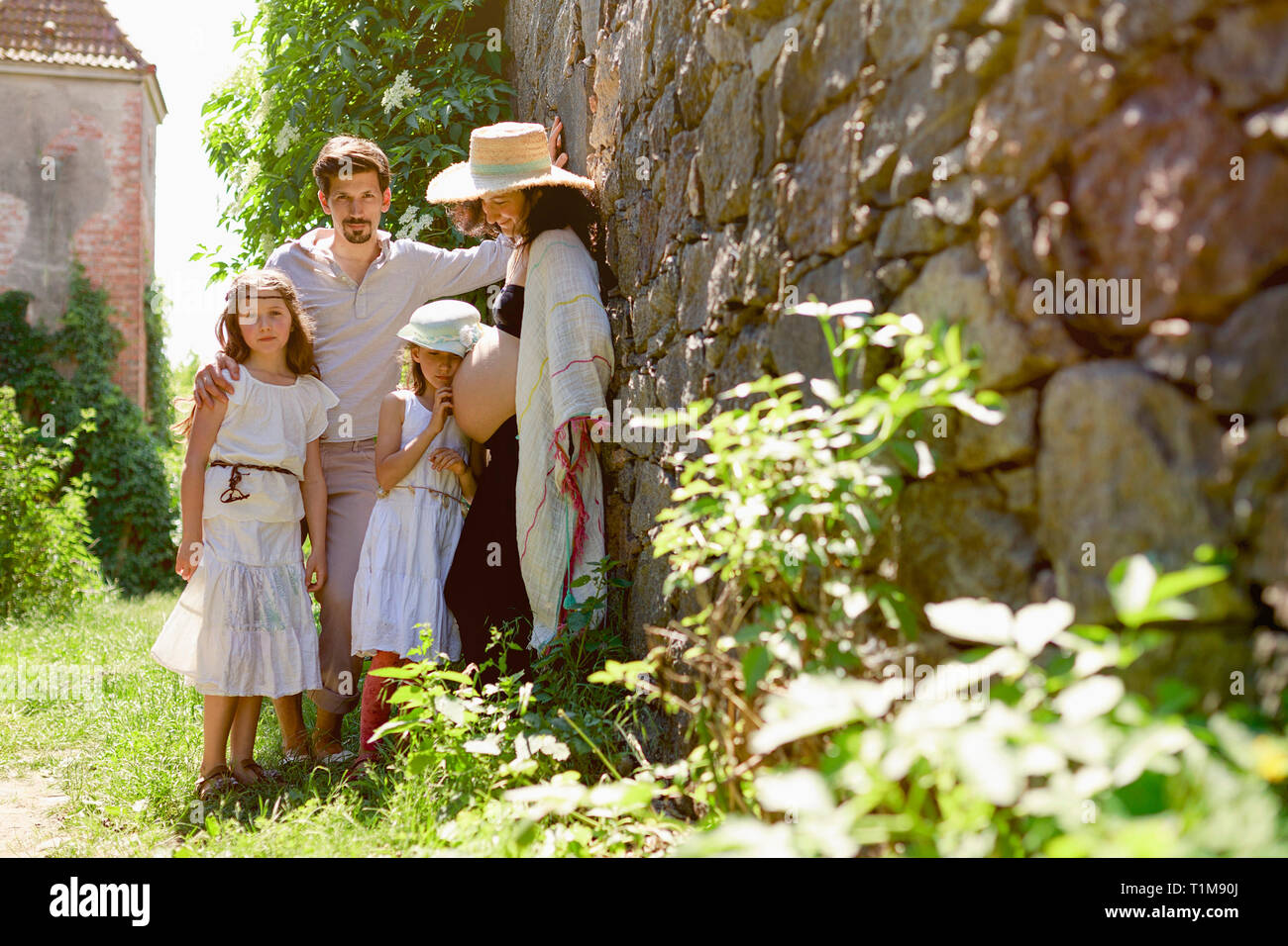Ritratto famiglia incinta al muro di pietra soleggiato in giardino Foto Stock