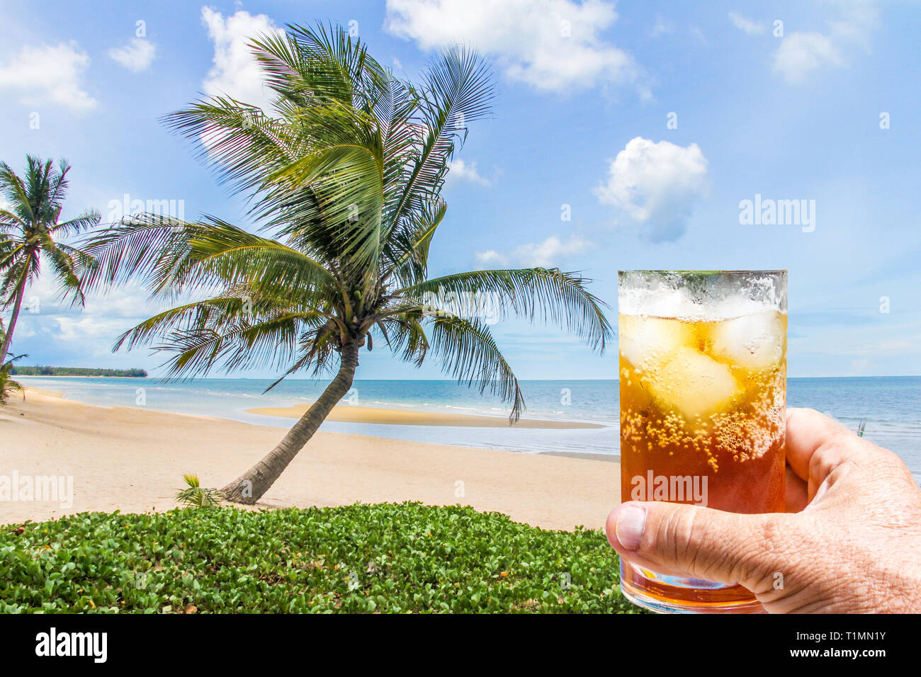 Mano che tiene un rum e coca cola cocktail nel bicchiere con ghiaccio sulla spiaggia con palme di cocco albero, Thailandia Foto Stock