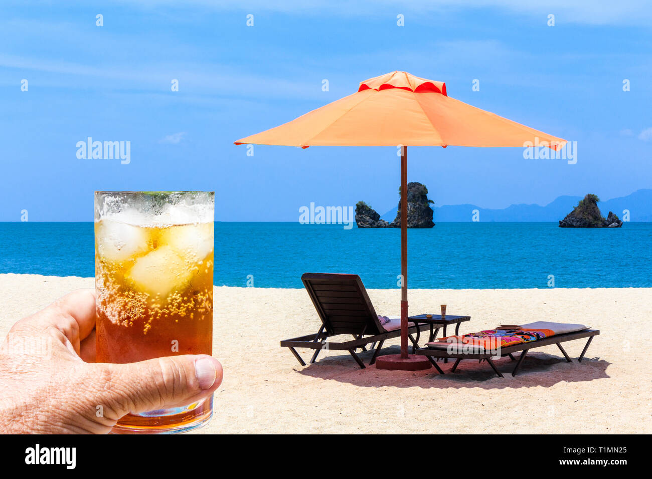 Mano che tiene un bicchiere di rum e coca cola con ghiaccio contro di sedie a sdraio e un ombrellone in un vuoto di Langkawi Beach, Malaysia Foto Stock