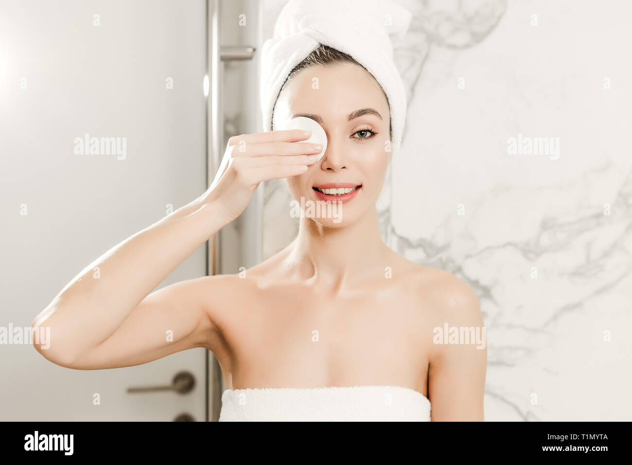 Giovane bella donna avvolta in asciugamani pulisce il suo volto con tampone di cotone in bagno. La bellezza trucco e concetto di cura della pelle Foto Stock