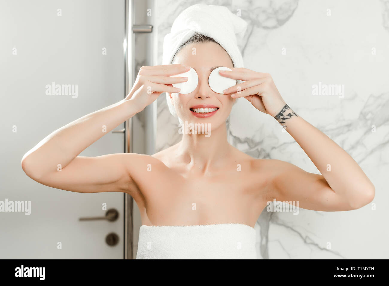 Giovane bella donna utilizza tamponi di cotone sui suoi occhi avvolto in asciugamani in bagno. Il trucco di bellezza e di cura della pelle concept Foto Stock