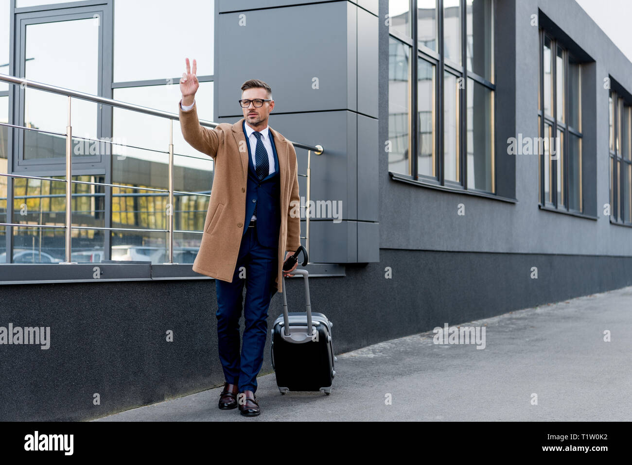 Bello imprenditore nel rivestire gesticolando mentre in piedi con la valigia e ombrello su strada Foto Stock