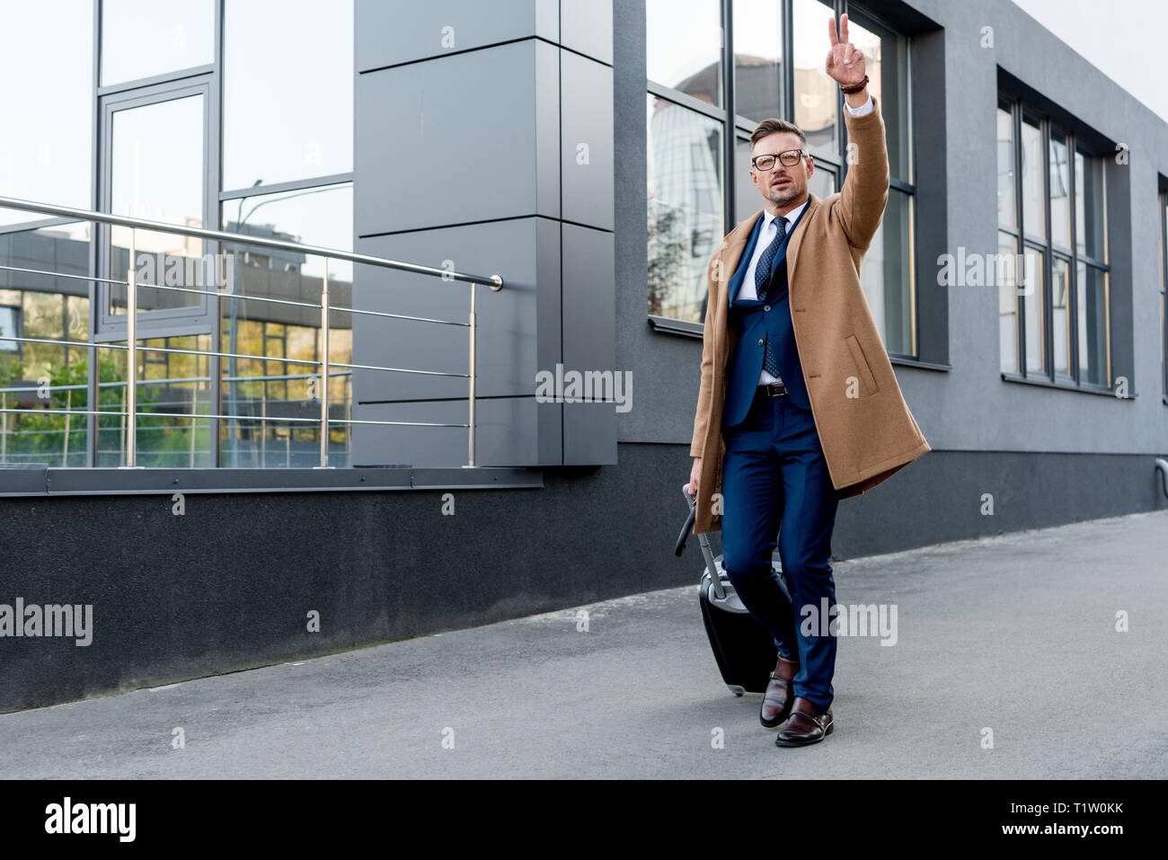 Imprenditore nel cappotto beige mostra segno di pace durante il funzionamento con la valigia e ombrello su strada Foto Stock