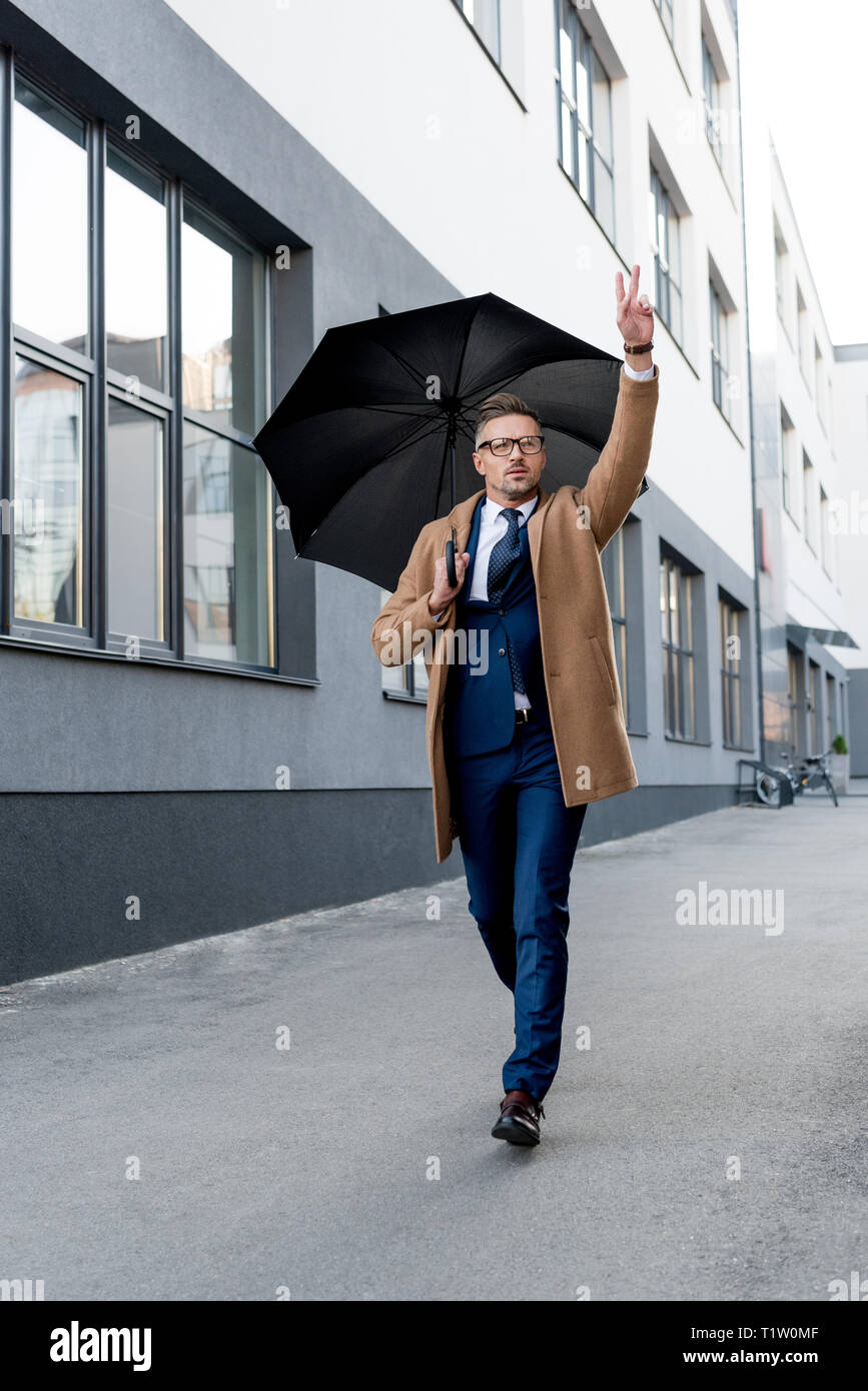 Bello imprenditore in bicchieri gesticolando mentre è in esecuzione la cappotto beige con ombrellone Foto Stock