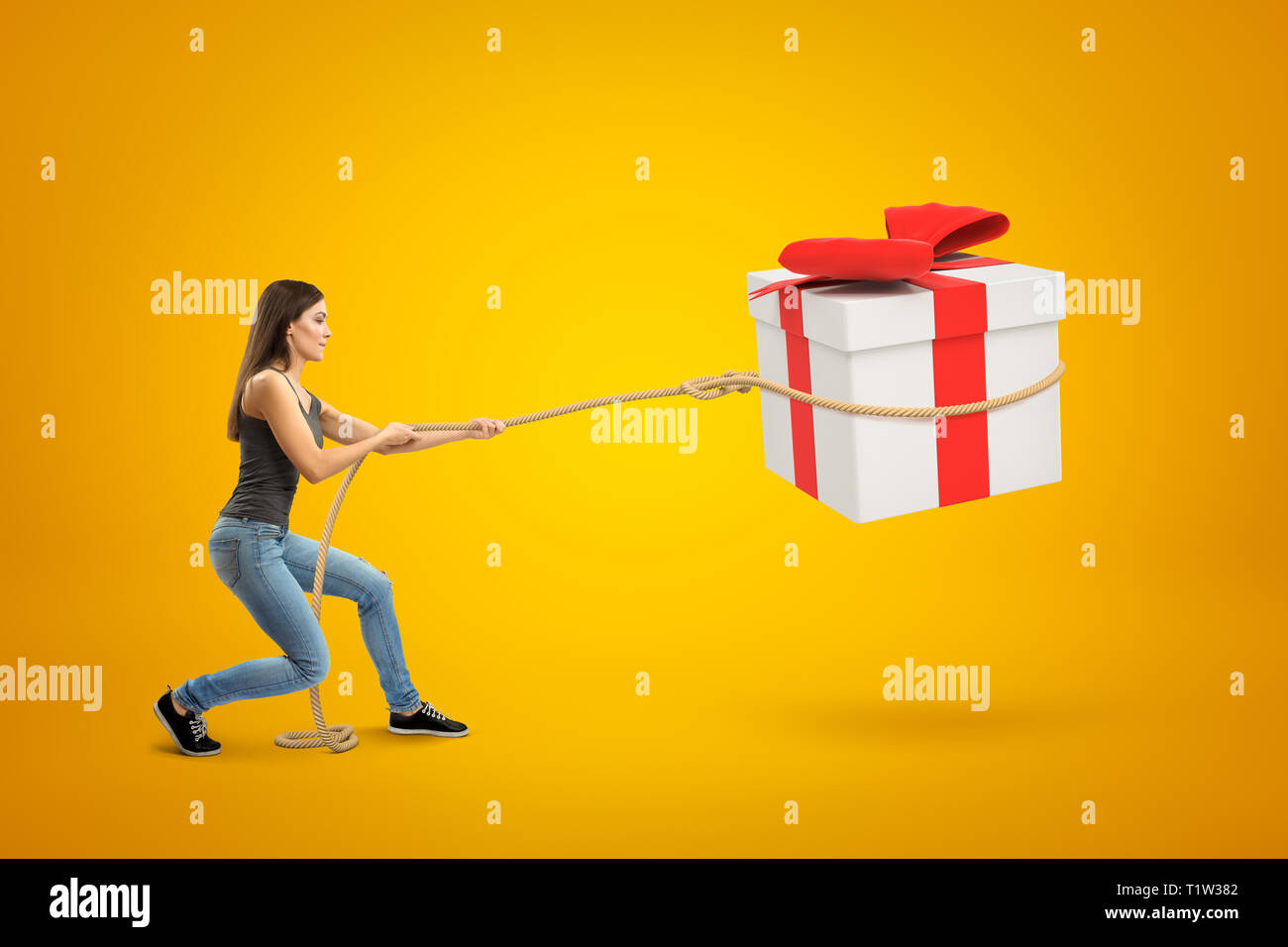 Vista laterale della giovane donna in piedi con le ginocchia piegate e tirando big box regalo in aria che ella ha lassoed, su sfondo giallo. Foto Stock