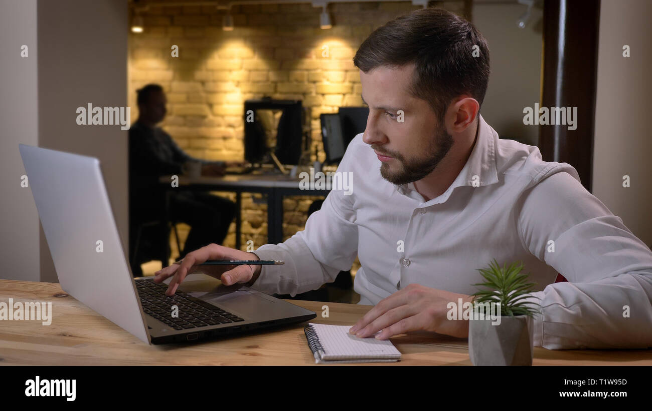 Closeup shoot di adulto imprenditore caucasico digitazione sul laptop e prendere appunti in ambienti chiusi in ufficio Foto Stock