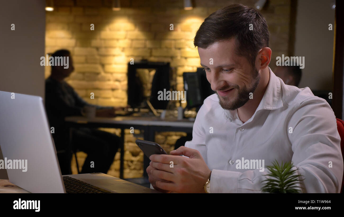Closeup shoot di adulto imprenditore caucasico utilizzando il telefono nella parte anteriore del computer portatile e sorridendo allegramente in ambienti chiusi in ufficio Foto Stock