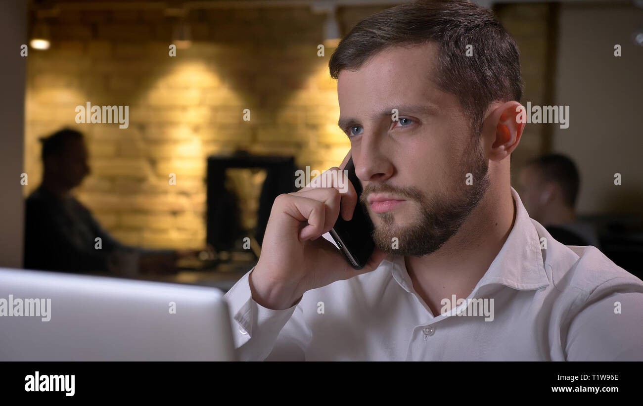 Closeup shoot di adulto imprenditore caucasico digitazione sul laptop e avere una formale chiamata telefonica in ambienti chiusi in ufficio Foto Stock