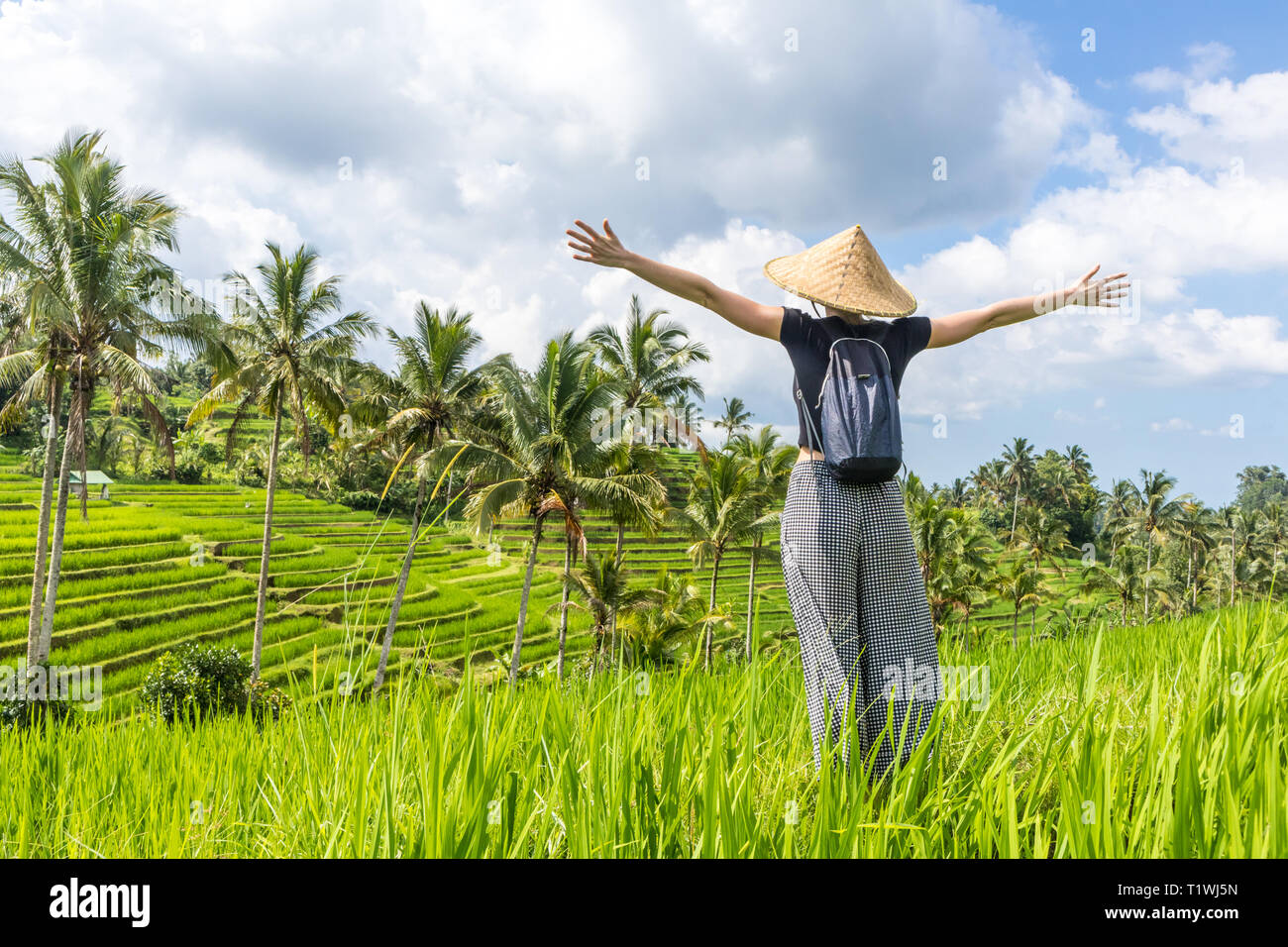 Alla moda rilassato viaggiatori femmina indossando il piccolo zaino e tradizionali asiatici paddy hat, bracci fatto alzare al cielo, godendo di natura pura in un bel verde Foto Stock