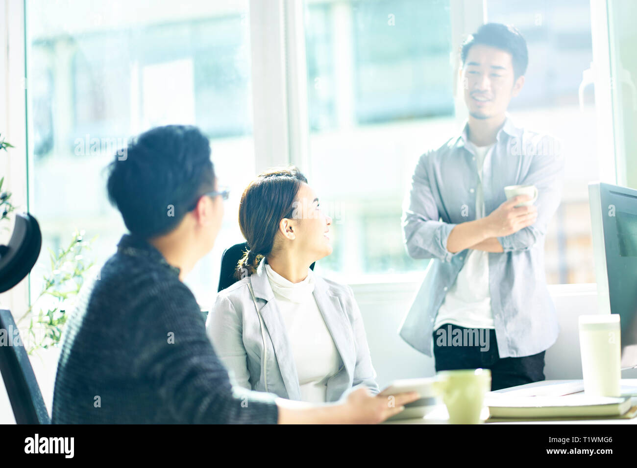 Tre giovani uomini asiatici e la donna in chat in ufficio, felici e rilassati. Foto Stock