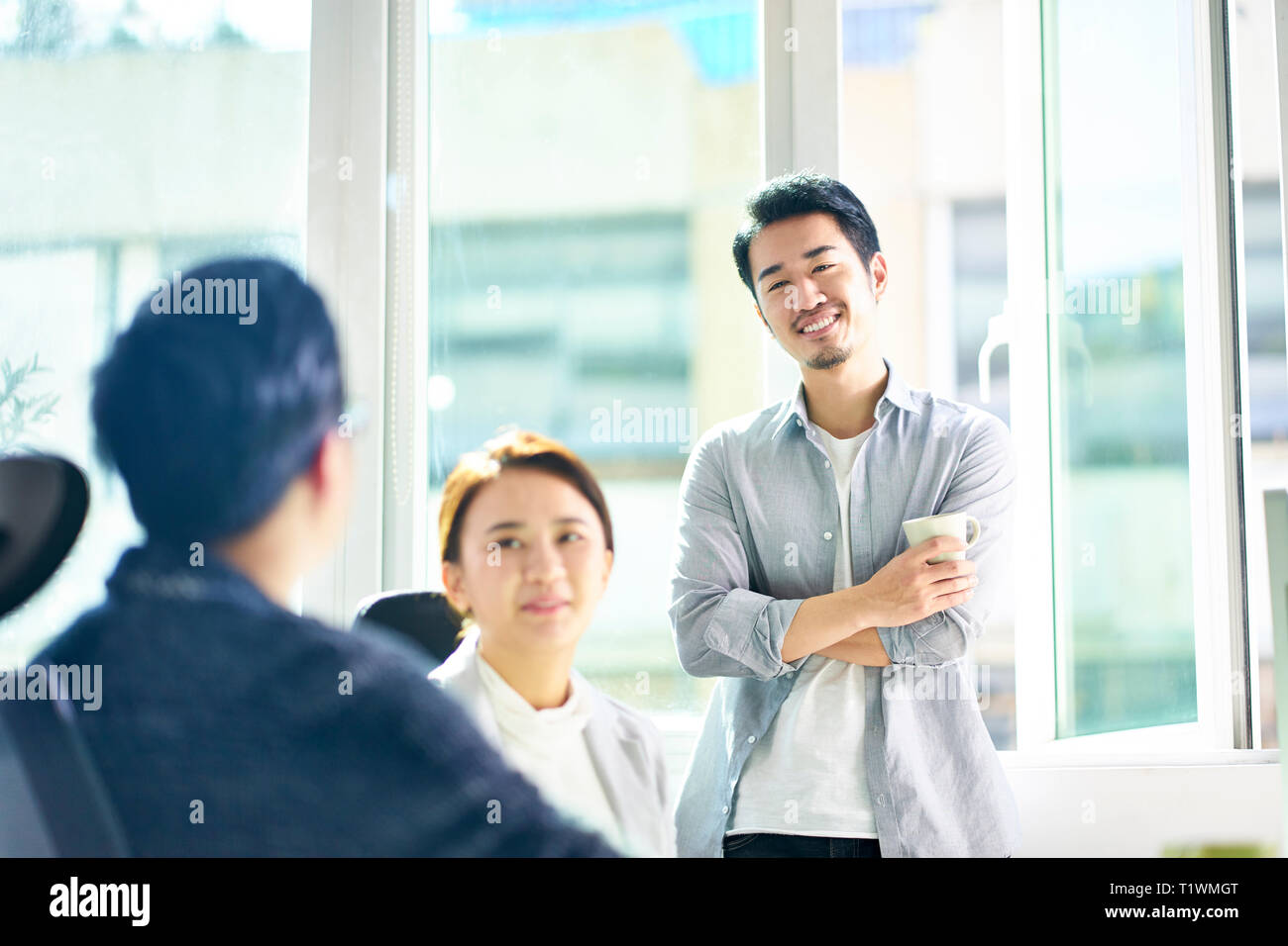 Tre giovani uomini asiatici e la donna in chat in ufficio, felici e rilassati. Foto Stock