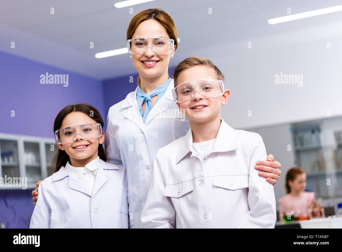 Maestro sorridente in camice bianco e occhiali protettivi abbracciando gli alunni durante la lezione di Chimica Foto Stock