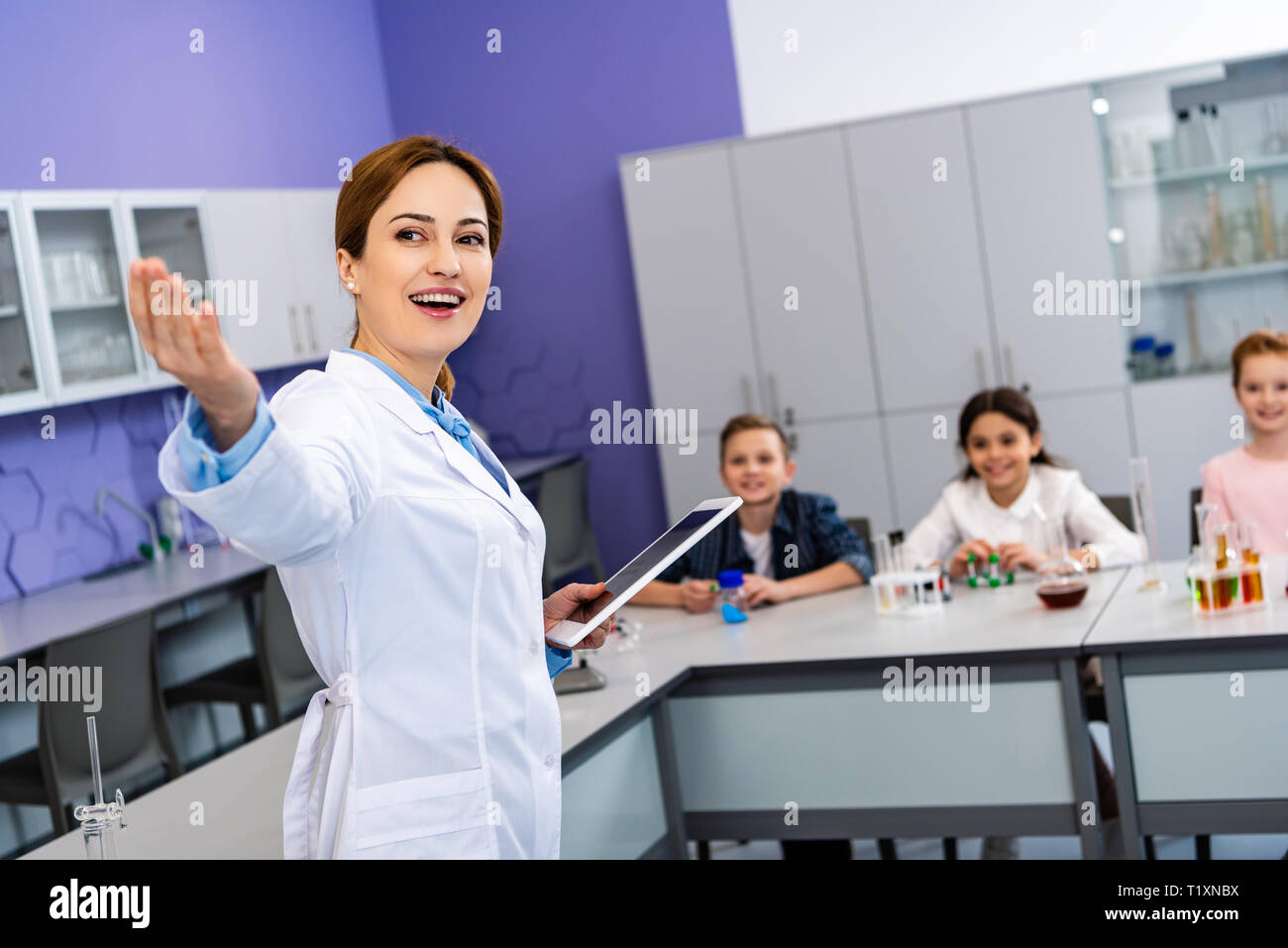 Sorridente insegnante di chimica in camice bianco azienda digitale compressa e indicando con la mano Foto Stock