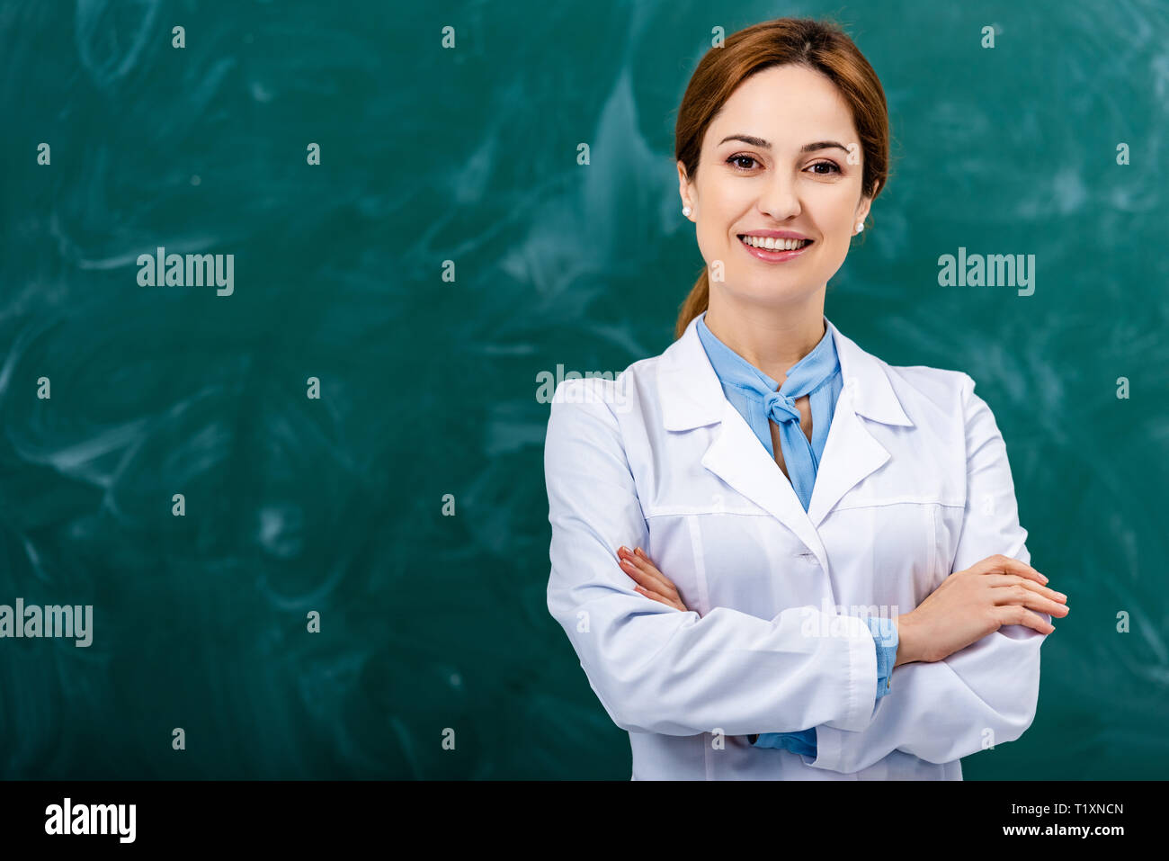 Sorridente insegnante di chimica in camice bianco in piedi con le braccia incrociate davanti di Blackboard Foto Stock